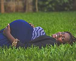 A pregnant woman laying in the grass smiling at the camera and holding her belly.