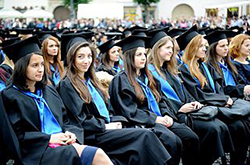 A row of female students sit at what appears to be a graduation ceremony. They each wear black robes and hats, with blue ties around their shoulders. One student looks at the camera, while the others have their gaze on the right side of the picture.
