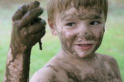 A kid with brown hair is covered in mud. We can see him from the shoulders up, and he is holding a dirty hand out in front of him. It looks like he has just crawled through the mud to find and hold up a worm.