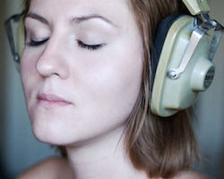 A girl is with medium length brown hair is wearing an old pair of headphones. She has her eyes closed and appears to be relaxing to the sound of the music