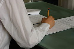 You see the arm of a person wearing a white dress shirt. They are sitting at a table holding a pencil. The paper they are working on is blurry, but it appears that they are reading through a test and filling in their answers on a scantron sheet.