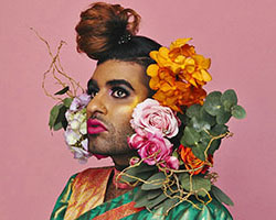 A portrait of Alok Vaid Menon surrounded by flowers against a pink background