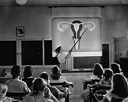 A black and white image of a teacher in 1950s style clothing pointing to a projected image of a uterus in front of the class.