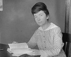 A black and white photo of Margaret Sanger sitting at a table in front of an open book while looking at the camera