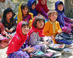 An image of young girls wearing head scarves sitting on the ground and smiling at something off camera.