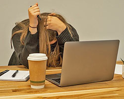A person with long hair sits in front of their laptop, a cup of coffee, and a notepad. They are looking down and clutching their head, feeling stress.