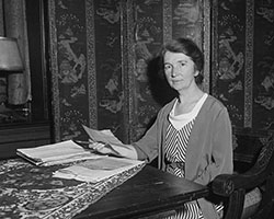 A black and white photo of Margaret Sanger sitting at a table holding a sheet of paper, looking at the camera.