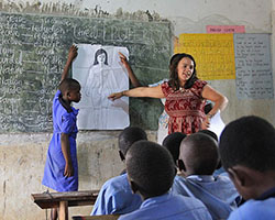 A student holds a drawing of the female body up against a chalkboard while a teacher points to the reproductive system.