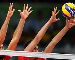 The hands of two people playing volleyball reach up, attempting to block a ball.