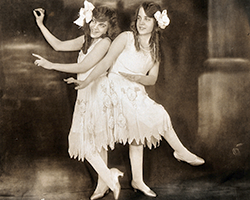 A pair of conjoined twins in dresses from the early 1900s dancing.