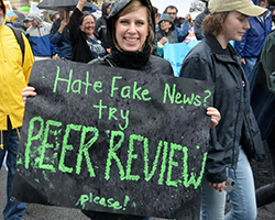 A woman holding a sign that says &quot;Hate fake news? Try peer review... please!&quot;