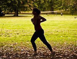 The image shows a person running in an open green area. The area has grass and trees in the background. The person is female, but is darkened by the shadow of the photo.