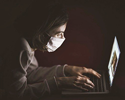 A girl at home, chatting with a friend on the computer, and wearing a mask.