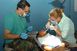 A male dentist (left) and a female assistant (right) work on a young patient who is reclined in a dental chair. Both of the dental workers wear teal colored scrubs. 