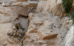 An archaeological dig site is shown, with the terrain being very sandy and rocky. The site shown in the picture is right next to a cliff face extending upwards to a height that cannot be seen. 