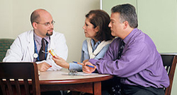a doctor, shown on the left, speaks with a middle-aged couple at a dark brown wooden table. The woman is wearing a blue shirt with a white sweater around her neck. THe man is wear a purple dress shirt and a dark colored tie.