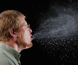 A man with red-brown hair and a beard is sneezing. The background is black, and you can see snot and other particles in the air as a result of his sneeze 
