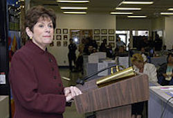 a woman wearing a maroon jacket stands at a podium ready to give a speech. You can see people behind her in the background of the image.